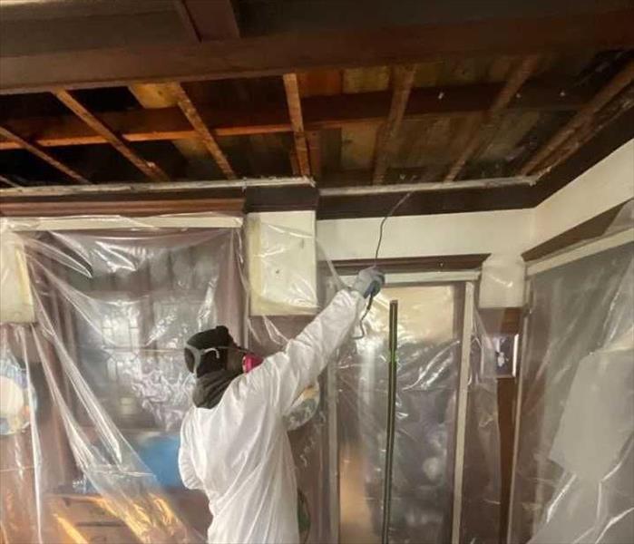 SERVPRO technician in full-body PPE in sealed and covered living room with open ceiling cavity.